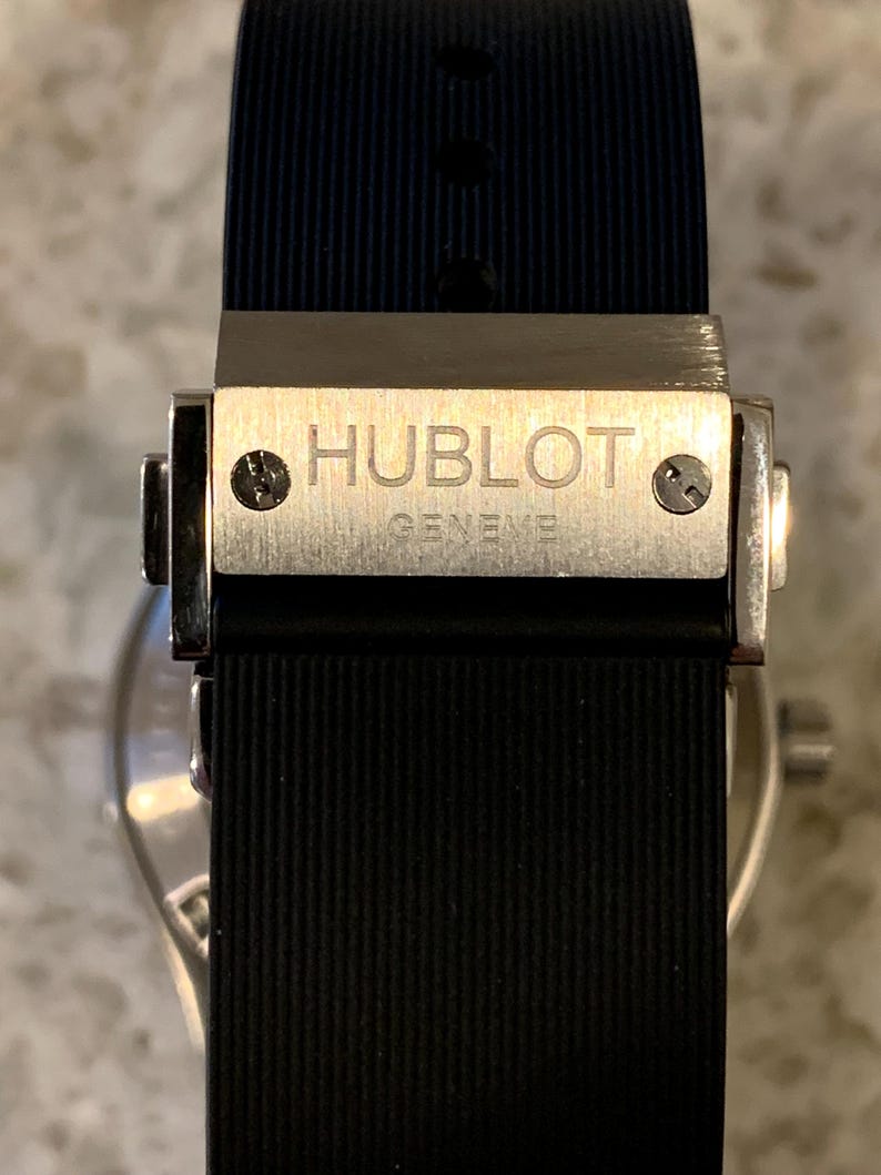 May include: Close-up of a luxury watch clasp. The silver clasp features the engraved brand name "HUBLOT GENEVE" and secures a black ribbed rubber strap. The watch band is dark blue on the inside.