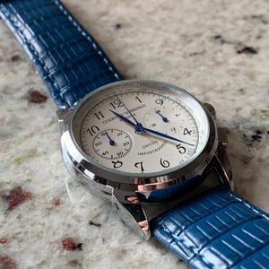 May include: A close-up of a wristwatch with a white face, silver casing, and blue hands. The watch has a blue leather band. The watch face displays the text "CUERVO Y SOBRINOS" and "UNICOS IMPORTADORES".