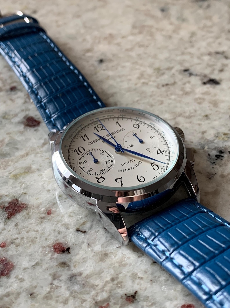 May include: A close-up of a wristwatch with a white face, silver casing, and blue hands. The watch has a blue leather band. The watch face displays the text "CUERVO Y SOBRINOS" and "UNICOS IMPORTADORES".