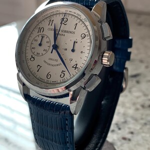 May include: A silver-toned wristwatch with a white face and blue hands. The watch features a blue leather band and multiple sub-dials. The watch face includes the text "CUERVO SOBRINOS HABANA" and "UNICOS IMPORTADORES".