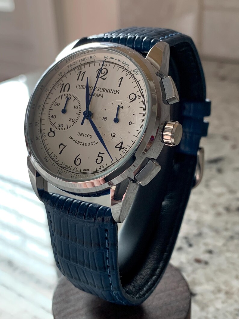 May include: A silver-toned wristwatch with a white face and blue hands. The watch features a blue leather band and multiple sub-dials. The watch face includes the text "CUERVO SOBRINOS HABANA" and "UNICOS IMPORTADORES".