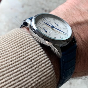May include: A silver wristwatch with a white face, blue hands, and a blue leather band. The watch has a round face with Arabic numerals and a subdial. The band has a textured, crocodile-like pattern.