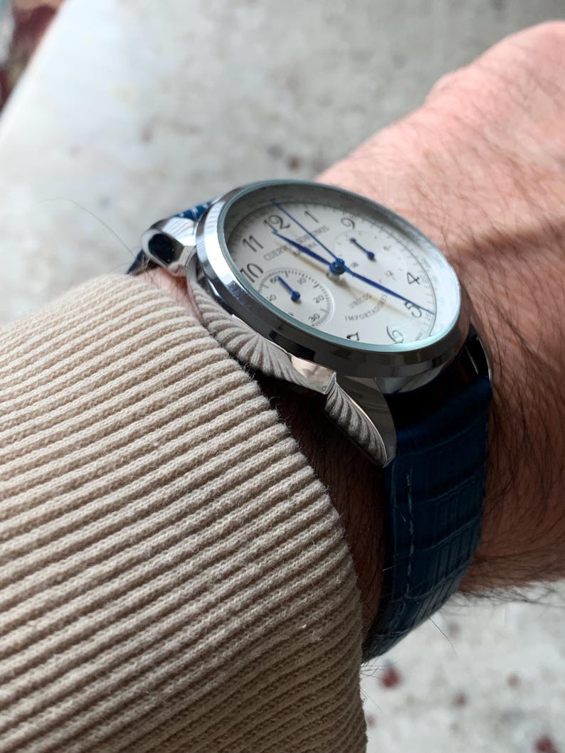 May include: A silver wristwatch with a white face, blue hands, and a blue leather band. The watch has a round face with Arabic numerals and a subdial. The band has a textured, crocodile-like pattern.