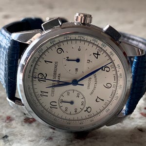 May include: A silver wristwatch with a white face, black numerals, and blue hands. The watch has a blue leather band. The face includes the text "CUERVO Y SOBRINOS HABANA UNICOS IMPORTADORE".