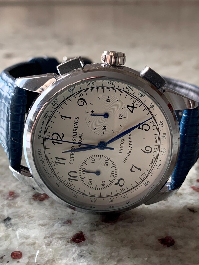 May include: A silver wristwatch with a white face, black numerals, and blue hands. The watch has a blue leather band. The face includes the text "CUERVO Y SOBRINOS HABANA UNICOS IMPORTADORE".