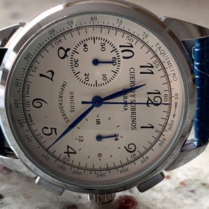 May include: A silver-toned wristwatch with a white face, featuring black numerals and blue hands. The watch has a blue leather strap and the text "CUERVO Y SOBRINOS" is visible on the face. The watch also has a tachymeter scale.