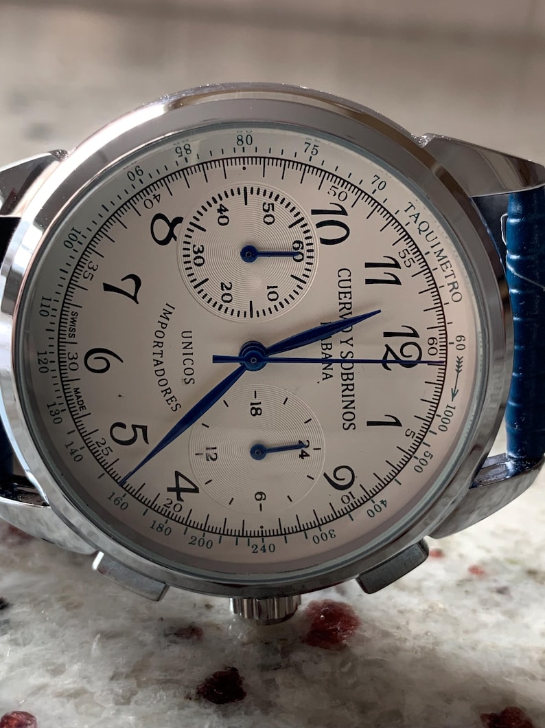 May include: A silver-toned wristwatch with a white face, featuring black numerals and blue hands. The watch has a blue leather strap and the text "CUERVO Y SOBRINOS" is visible on the face. The watch also has a tachymeter scale.