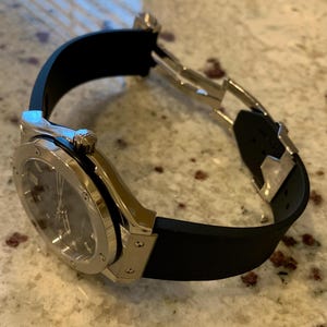 May include: A luxury wristwatch with a silver-colored case and a black rubber strap. The watch face is dark gray. The watch is angled, showing the clasp and the details of the watch's design.