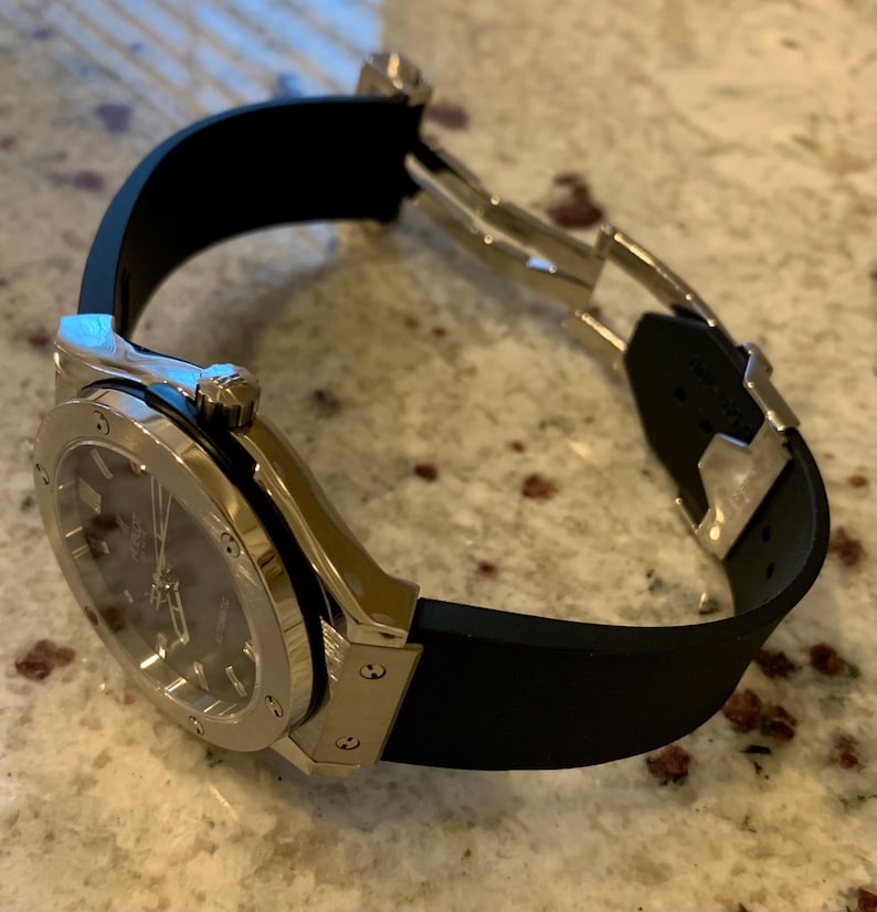 May include: A luxury wristwatch with a silver-colored case and a black rubber strap. The watch face is dark gray. The watch is angled, showing the clasp and the details of the watch's design.