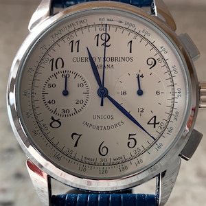 May include: A silver wristwatch with a white face, black numerals, and blue hands. The watch features a blue leather band and the text "CUERVO Y SOBRINOS HABANA" and "UNICOS IMPORTADORES". The watch has a tachymeter scale.