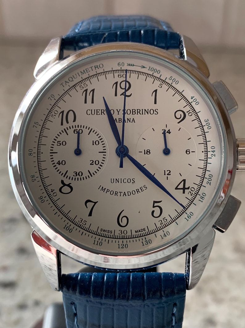 May include: A silver wristwatch with a white face, black numerals, and blue hands. The watch features a blue leather band and the text "CUERVO Y SOBRINOS HABANA" and "UNICOS IMPORTADORES". The watch has a tachymeter scale.