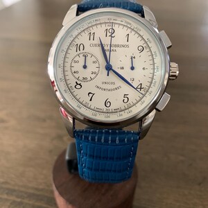 May include: A silver-toned wristwatch with a white face, featuring black numerals and blue hands. The watch has a blue leather band and a tachymeter scale. The watch face displays the text "CUERVO Y SOBRINOS HABANA" and "UNICOS IMPORTADORES".