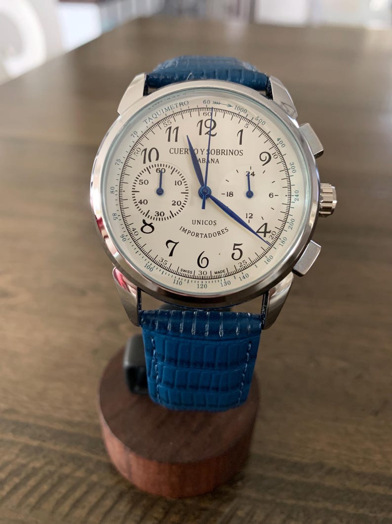 May include: A silver-toned wristwatch with a white face, featuring black numerals and blue hands. The watch has a blue leather band and a tachymeter scale. The watch face displays the text "CUERVO Y SOBRINOS HABANA" and "UNICOS IMPORTADORES".
