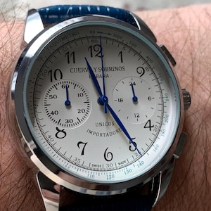 May include: A silver-toned wristwatch with a white face, black numerals, and blue hands. The watch features multiple sub-dials and a blue leather band. The watch face displays the brand name "CUERVO Y SOBRINOS" and the word "UNICOS".