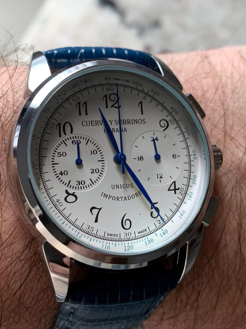 May include: A silver-toned wristwatch with a white face, black numerals, and blue hands. The watch features multiple sub-dials and a blue leather band. The watch face displays the brand name "CUERVO Y SOBRINOS" and the word "UNICOS".
