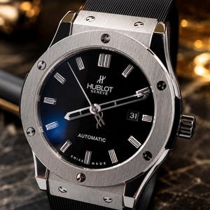 May include: A luxury wristwatch with a black face and silver-tone bezel. The watch features the brand name "Hublot Geneve" and the word "Automatic". The watch has a black rubber strap and a date display at the 3 o'clock position.