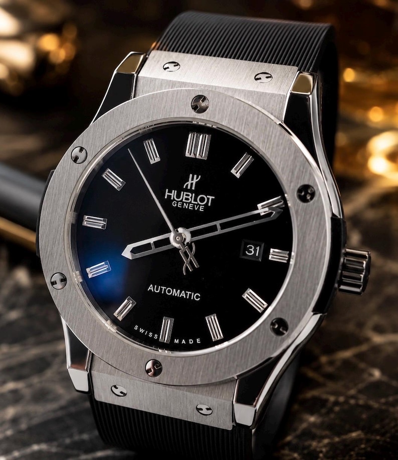 May include: A luxury wristwatch with a black face and silver-tone bezel. The watch features the brand name "Hublot Geneve" and the word "Automatic". The watch has a black rubber strap and a date display at the 3 o'clock position.