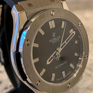 May include: A stainless steel Hublot Geneve automatic watch with a black dial. The watch features silver hands and hour markers, a date window, and a black rubber strap. The watch is displayed on a black stand.