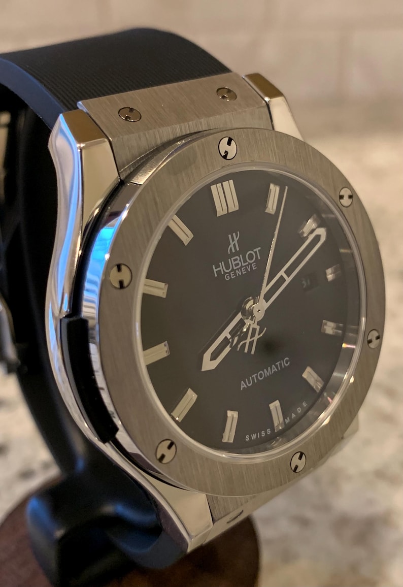 May include: A stainless steel Hublot Geneve automatic watch with a black dial. The watch features silver hands and hour markers, a date window, and a black rubber strap. The watch is displayed on a black stand.
