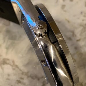 May include: Close-up of a silver wristwatch with a black band. The watch has a round face and a textured crown. The watch is made of polished metal and has a sleek, modern design. The watch is a luxury item.