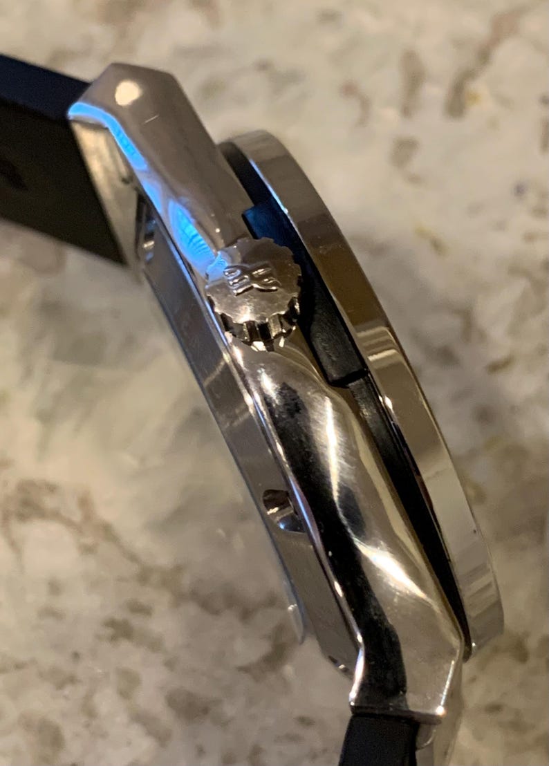 May include: Close-up of a silver wristwatch with a black band. The watch has a round face and a textured crown. The watch is made of polished metal and has a sleek, modern design. The watch is a luxury item.