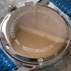 May include: Close-up of a silver-toned watch with a blue leather strap. The watch face features the engraved text "CUERVO Y SOBRINOS - HABANA" and "HISTORIADOR". The watch has a round face and a textured, reflective surface.