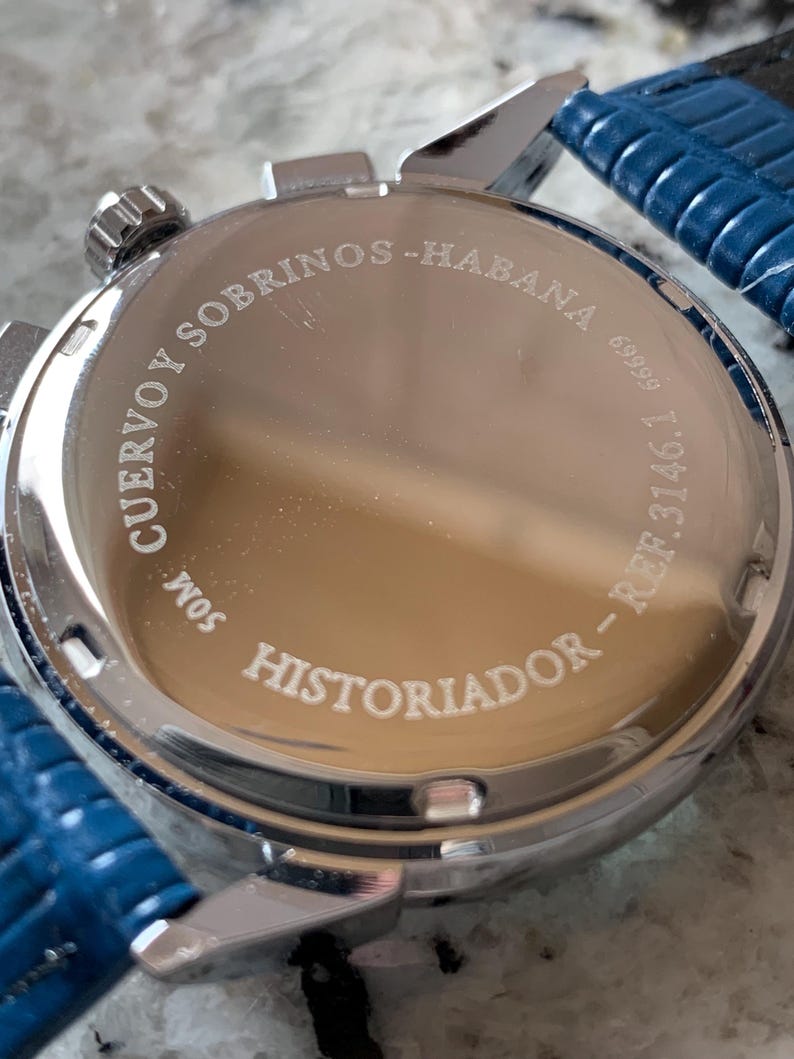 May include: Close-up of a silver-toned watch with a blue leather strap. The watch face features the engraved text "CUERVO Y SOBRINOS - HABANA" and "HISTORIADOR". The watch has a round face and a textured, reflective surface.