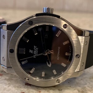 May include: A stainless steel Hublot automatic watch with a black dial. The watch features silver-tone hour markers and hands, a date display at the 3 o'clock position, and a black rubber strap. The watch face has the text "Hublot Geneve Automatic Swiss Made".