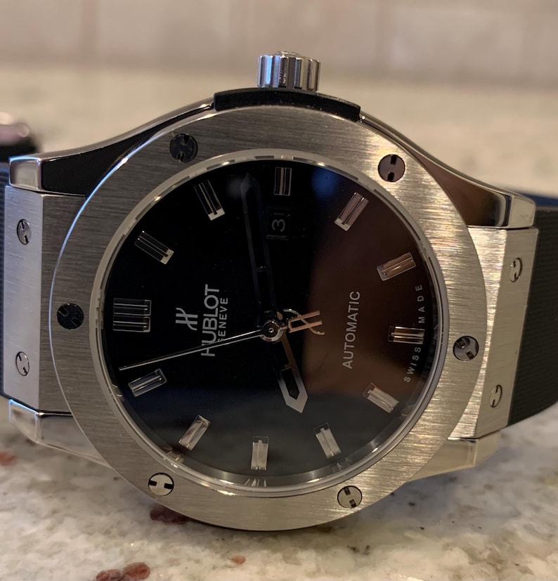 May include: A stainless steel Hublot automatic watch with a black dial. The watch features silver-tone hour markers and hands, a date display at the 3 o'clock position, and a black rubber strap. The watch face has the text "Hublot Geneve Automatic Swiss Made".