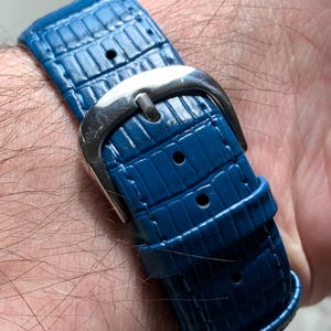 May include: Close-up of a blue watch strap with a silver buckle. The strap has a textured, reptile-like pattern. The buckle is a polished silver color. The watch strap is secured on a wrist.