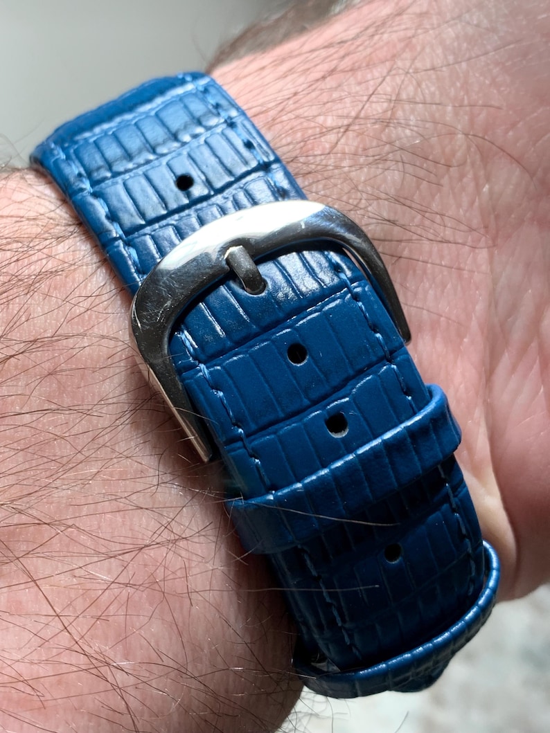 May include: Close-up of a blue watch strap with a silver buckle. The strap has a textured, reptile-like pattern. The buckle is a polished silver color. The watch strap is secured on a wrist.
