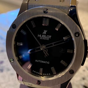 May include: A luxury wristwatch with a black face and silver-tone bezel. The watch features the brand name "Hublot Geneve" and the word "Automatic". The watch has a black band and is displayed on a wooden stand.