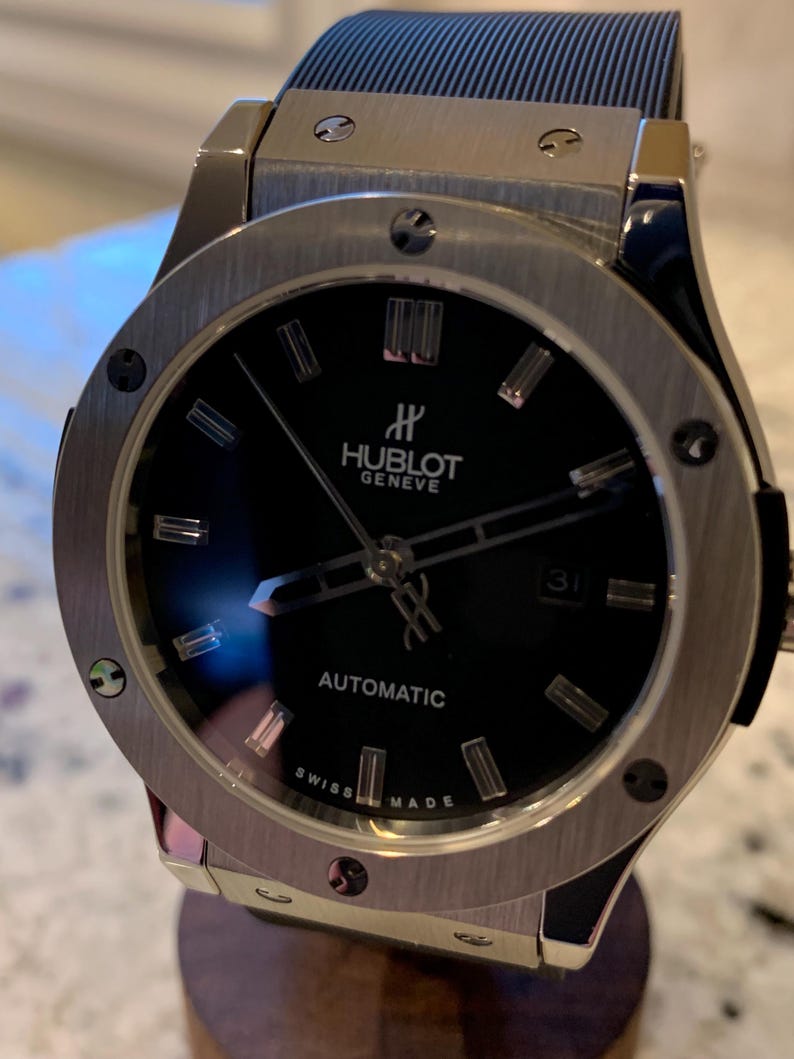 May include: A luxury wristwatch with a black face and silver-tone bezel. The watch features the brand name "Hublot Geneve" and the word "Automatic". The watch has a black band and is displayed on a wooden stand.