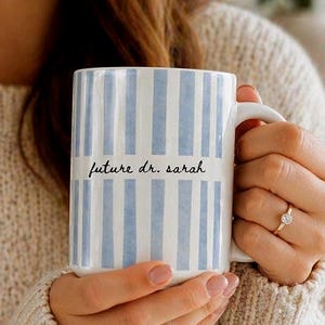 May include: A white ceramic mug with vertical light blue stripes. The mug has the text "future dr. sarah" in black script. The mug is held by a person wearing a beige sweater.