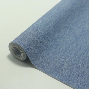 May include: A roll of blue denim-textured wallpaper. The wallpaper has a light blue color with a subtle, textured pattern, mimicking the look of denim fabric. The roll is partially unrolled, revealing the paper's core.