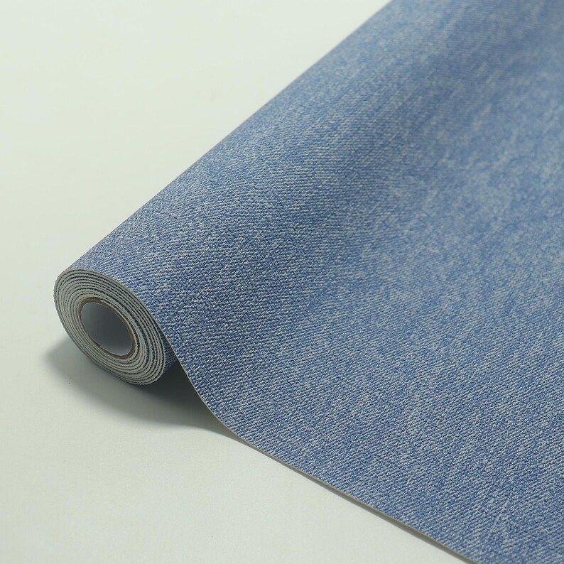 May include: A roll of blue denim-textured wallpaper. The wallpaper has a light blue color with a subtle, textured pattern, mimicking the look of denim fabric. The roll is partially unrolled, revealing the paper's core.