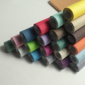 Op de afbeelding: Een verzameling opgerolde stoffen in verschillende kleuren, waaronder rood, blauw, groen en geel. De rollen zijn opgestapeld en tonen de verschillende tinten en texturen van het materiaal. De stof lijkt geweven.