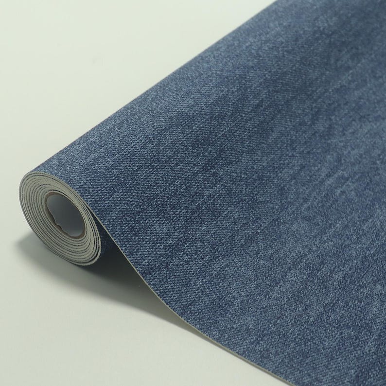 May include: A roll of blue denim-textured wallpaper. The wallpaper has a woven appearance, with variations in the blue color creating a realistic denim look. The roll is partially unrolled, revealing the white backing.