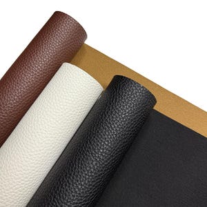 May include: Close-up of rolled faux leather sheets in various colours. The rolls include brown, white, black, and tan. The material has a textured, pebbled surface.