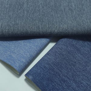 May include: Close-up of three pieces of denim fabric in varying shades of blue. The top piece is a darker, muted blue, while the lower pieces are lighter shades of blue. The fabric has a textured appearance, with visible weave details.
