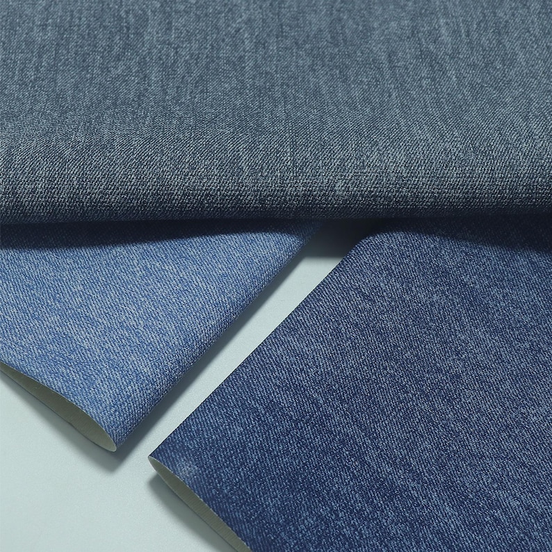 May include: Close-up of three pieces of denim fabric in varying shades of blue. The top piece is a darker, muted blue, while the lower pieces are lighter shades of blue. The fabric has a textured appearance, with visible weave details.