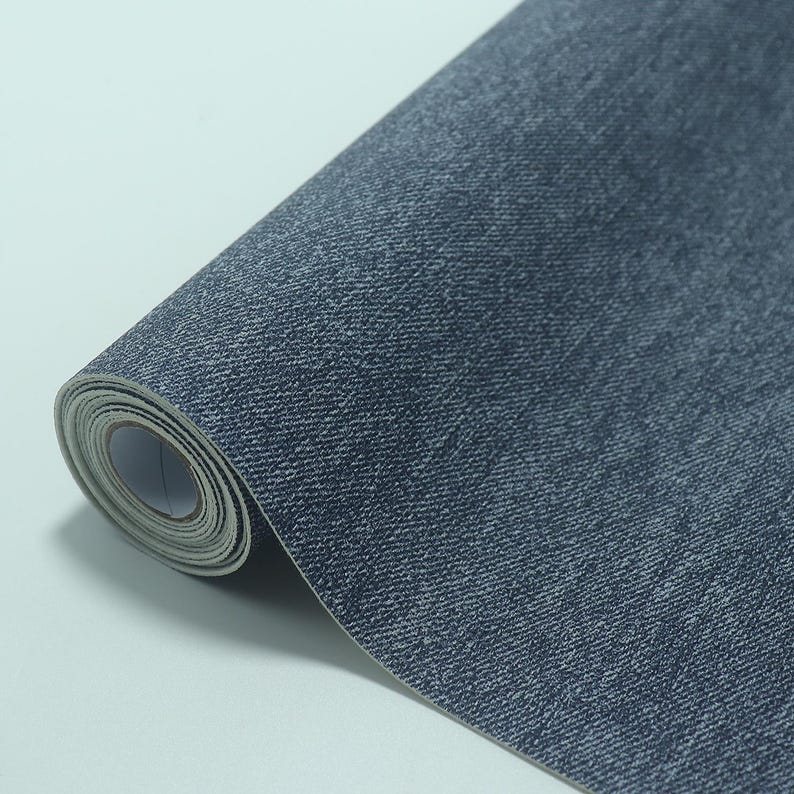 May include: A roll of denim-textured wallpaper. The wallpaper is a dark blue color with a subtle, woven pattern. The roll is partially unrolled, revealing the paper's texture and the white backing.