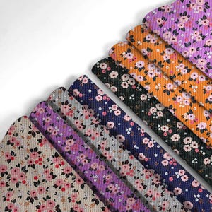 以下が含まれることがあります： ベージュ、紫、グレー、ネイビー、オレンジ、黒など、さまざまな色の花柄生地の見本。各見本は、緑の葉を持つピンクと白の花の繰り返し模様を特徴としています。生地は斜めの扇形に配置されています。