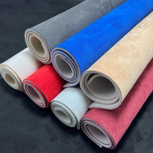 Può includere: Una collezione di tessuto scamosciato arrotolato in vari colori, tra cui grigio, blu, beige e rosso. I rotoli sono impilati, mostrando le diverse tonalità e trame del materiale. La pelle scamosciata appare morbida e ha una superficie liscia.