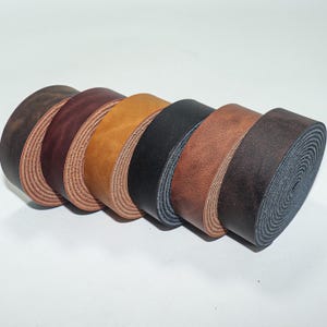 Peut inclure: Plusieurs bandes de cuir roulées de différentes couleurs : marron foncé, bordeaux, beige, noir et marron clair. Les bords du cuir présentent une teinte claire et naturelle. Les rouleaux sont disposés en rangée sur fond blanc.