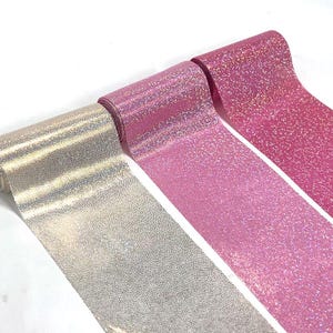 May include: Three rolls of decorative ribbon in silver, light pink, and hot pink. The ribbon has a textured, glittery surface, and is unrolled to show the width. The rolls are set against a white background.