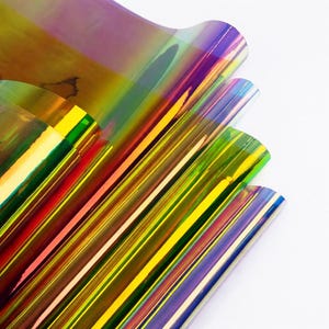 Op de afbeelding: Close-up van iriserende filmvellen in tinten geel, groen, paars en rood. De vellen zijn gelaagd en schuin geplaatst, waardoor een gestreept patroon ontstaat. Het materiaal heeft een glanzend, reflecterend oppervlak.