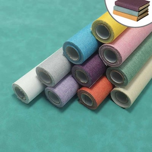 May include: A collection of ten fabric rolls in various solid colors including white, gray, blue, yellow, pink, purple, green, orange, and cream, arranged in a pyramid formation, with an inset showing three stacked books in purple, blue, and orange.