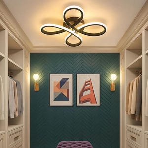May include: A contemporary walk-in wardrobe with white cabinets, a teal feature wall, and a purple footstool. The ceiling has a distinctive, looping light fitting. Two framed pictures adorn the wall, and two sconces offer extra illumination.