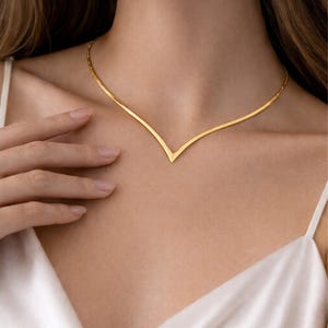 Könnte beinhalten: Eine zarte goldfarbene Halskette mit einem V-förmigen Anhänger. Die Halskette wird am Hals einer Person getragen, und das Design ist schlicht und elegant. Die Halskette besteht aus einem dünnen, glänzenden Metall.